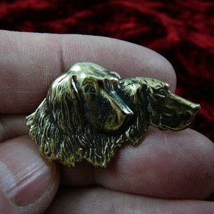 Op de afbeelding: Een bronzen metalen pin met twee hondenkoppen, een bovenop de andere. De honden kijken naar rechts, met hun oren en neuzen zichtbaar. De pin is gedetailleerd en realistisch.