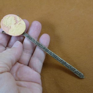 May include: A bronze bookmark with an ornate design and a cameo of a woman's profile in pink and white.