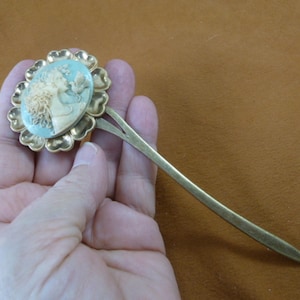 May include: A gold hair stick with a floral design. The flower has a cameo of a woman's face in the center. The stick is about 7 inches long.