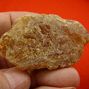 May include: A raw, unpolished piece of amber-colored resin with a translucent, crystalline texture. The stone features irregular, jagged edges and a warm, golden-brown hue with varying levels of opacity across its uneven, weathered surface.