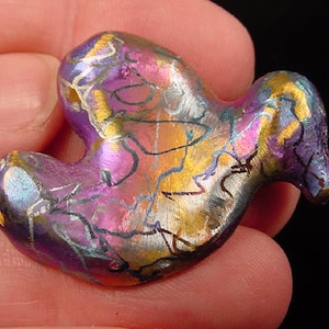 Puede incluir: Una pequeña escultura de pájaro de cerámica abstracta con un esmalte iridiscente morado, dorado y plateado. El pájaro tiene un cuerpo redondeado y un pico largo y delgado.