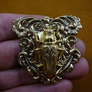 May include: A gold-colored brooch featuring a detailed insect design at the center, surrounded by ornate, swirling embellishments. The brooch has a vintage aesthetic and is likely made of metal.