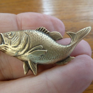 May include: A gold-coloured metal fish figurine with detailed scales and fins. The fish has its mouth open and its tail is curled up.