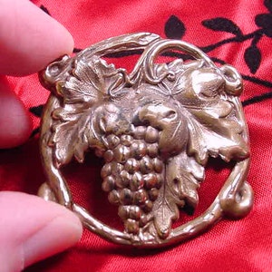May include: A gold-toned metal brooch in the shape of a grapevine with grapes. The grapes are clustered together and the leaves are detailed.