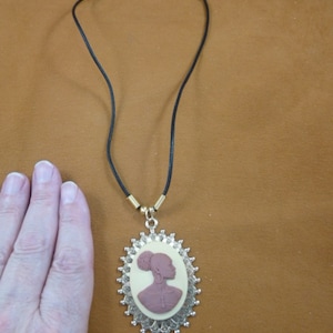 Puede incluir: Un collar de cordón negro con un colgante de camafeo ovalado dorado que presenta una silueta marrón de una mujer con un moño. El camafeo está rodeado por un diseño de filigrana dorada.
