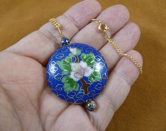 Könnte beinhalten: Eine blaue und goldene Cloisonné-Halskette mit einem Blumenmuster. Der Anhänger zeigt eine weiße Blume mit rosa Akzenten und grünen Blättern auf blauem Hintergrund. Die Kette besteht aus einer goldenen Kette mit einem Verschluss.