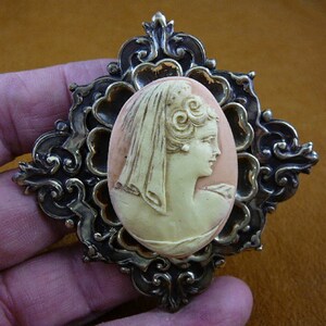 May include: A cameo brooch with a detailed ornate frame in gold and silver tones. The cameo features a woman's profile in white and pink.