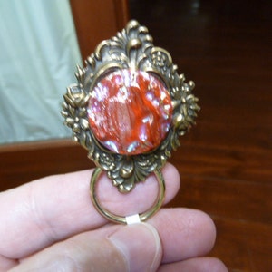 Puede incluir: Un cabujón de vidrio rojo e iridiscente engastado en un marco de metal dorado adornado con un anillo adjunto.