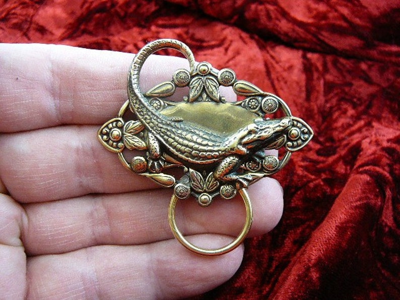 May include: A gold-toned alligator brooch with intricate details. The alligator is curled up with its tail looped around its body. The brooch has a decorative design with leaves and flowers.