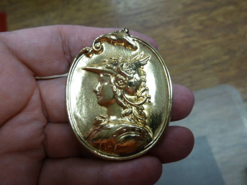 May include: A gold-toned pendant featuring a detailed relief of a woman's profile wearing a helmet with a feathered crest. The pendant is oval-shaped and has a smooth, polished surface.
