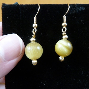 Może przedstawiać: Złote kolczyki z okrągłym, jasnożółtym koralikiem z tygrysiego oka na każdym kolczyku.