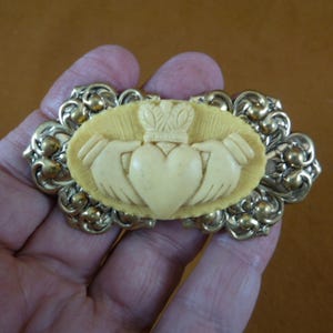 May include: A gold-toned brooch with a carved ivory-colored design of two hands clasped around a heart with a crown on top.