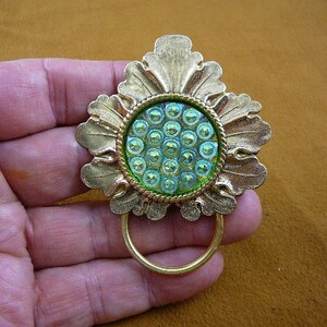 Puede incluir: Un broche de metal dorado en forma de flor con un centro de vidrio verde iridiscente. El centro está decorado con un patrón de pequeños círculos en relieve.