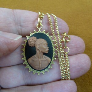 May include: Gold chain necklace with a cameo pendant featuring a brown sculpted profile of a woman with a bun hairstyle. The cameo is set in a gold-toned oval frame with a black background.