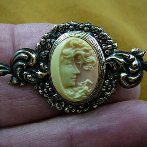 May include: Vintage cameo brooch featuring a profile of a woman with flowing hair. The oval cameo is set in a gold-toned metal frame with ornate scrollwork and floral accents. The cameo is a light peach color, and the frame has a dark, antique finish.
