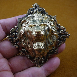 Può includere: Una spilla a forma di testa di leone dorata con una cornice ornata dettagliata. La testa di leone è tridimensionale e ha un'espressione feroce.