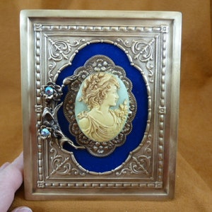 May include: A gold-toned metal box with an ornate frame and a blue velvet interior. The box features a cameo of a woman's profile with a floral design. The cameo is surrounded by a gold-toned metal frame with a floral design. The box has two decorative floral accents with blue gemstones.