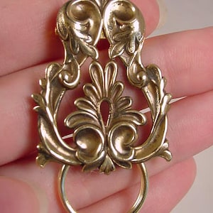 May include: A gold-toned metal brooch with an intricate, ornate design featuring a ring at the bottom. The brooch has a floral motif and a delicate, filigree style.