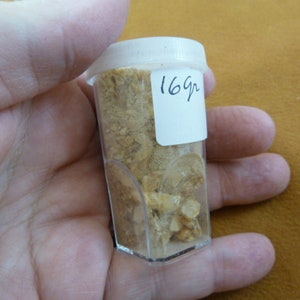 May include: A clear plastic container filled with a light brown substance. A white label on the container reads "16g".