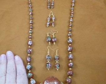Puede incluir: Un collar marrón y dorado con un gran colgante redondo y pendientes a juego. El collar y los pendientes están hechos de cuentas con un diseño floral.