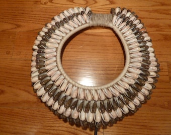 May include: A large, round necklace made of many small, white and brown seashells. The shells are arranged in a layered pattern, creating a textured and intricate design. The necklace is tied together with a thick, natural fiber cord.
