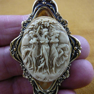 Puede incluir: Un broche de camafeo con un engaste dorado presenta un diseño tallado de tres mujeres de blanco, sosteniendo una corona de flores. El camafeo está engastado en un marco ovalado decorativo con un acabado negro y dorado.