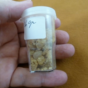 May include: A small, clear plastic container with a white lid, filled with small, light-colored, irregularly shaped stones. A white label on the container reads "16 gr". The container is held in a hand against a brown background.
