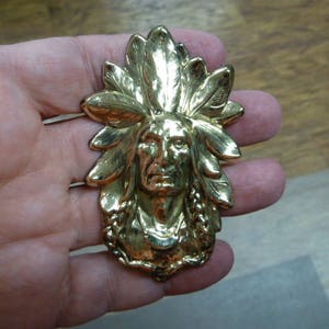 May include: A gold-colored metal pendant depicting a Native American man wearing a headdress. The pendant is detailed with feathers and beads.