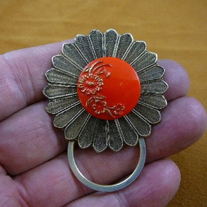Puede incluir: Un broche de metal plateado en forma de flor con un centro de esmalte rojo y detalles florales dorados. El broche tiene un bucle en la parte posterior para sujetarlo a la ropa.
