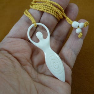 Puede incluir: Collar con colgante de hueso blanco tallado con un diseño en espiral y un cordón amarillo. El colgante tiene la forma de una figura humana estilizada.
