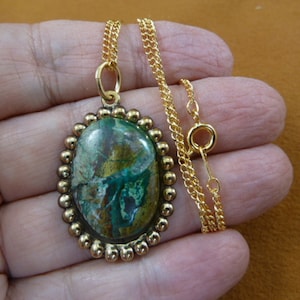 May include: Gold chain necklace with a green and brown oval pendant. The pendant is set in a gold bezel with a beaded edge.