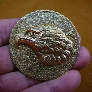May include: A gold-toned metal eagle head pendant with intricate filigree details. The eagle's head is facing right and has a detailed beak and feathers.