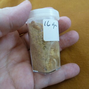 May include: A clear plastic container with a white label that says "16 gr" filled with a light brown powder.