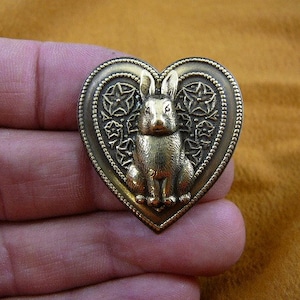 May include: A gold-toned heart-shaped pin with a detailed design featuring a rabbit in the center. The rabbit is sitting upright with its ears perked up.