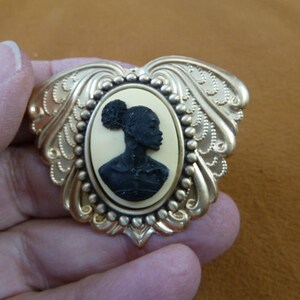 May include: Gold tone brooch with a cameo featuring a black silhouette of a woman with a bun in her hair. The cameo is set in a white oval.