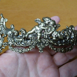 Könnte beinhalten: Eine goldfarbene Haarklammer aus Metall mit einem dekorativen Design, das zwei Cherubim zeigt, die eine Blume halten. Die Klammer hat ein detailliertes, verziertes Design mit filigranen Schnörkeln und floralen Akzenten.