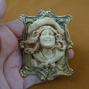 May include: A vintage-style brooch featuring a woman's face in a cameo-like design. The woman is wearing a hat and has long, flowing hair. The cameo is set in a gold-toned frame with intricate details.