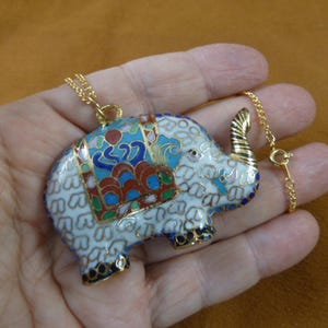 May include: A white enamel elephant pendant necklace with blue, red, and green details. The elephant has a gold chain and a gold clasp.
