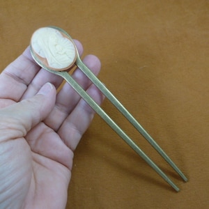May include: A pair of gold-toned hair sticks with a carved oval cameo at the top. The cameo is white with a raised design and a copper-toned border.