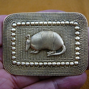 May include: Gold-toned metal belt buckle with a raised, detailed armadillo design. The buckle has a textured, patterned border.