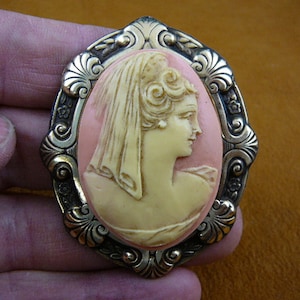 May include: A cameo brooch with a detailed carving of a woman's profile wearing a veil. The cameo is set in a gold-toned metal frame with an ornate design.