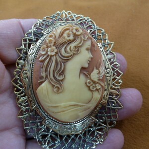 May include: A gold-tone cameo brooch with a detailed filigree design. The cameo features a woman's profile with a bird and flowers.