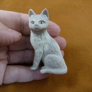 Könnte beinhalten: Eine handgeschnitzte weiße Katzenfigur, die mit ihrem Schwanz um die Beine gewickelt sitzt. Die Figur ist aus einem hellen Material, möglicherweise Knochen oder Elfenbein, gefertigt.
