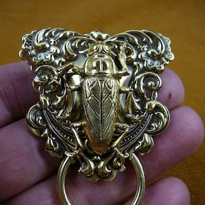 May include: A gold-toned metal brooch featuring a detailed beetle design with intricate floral accents. The brooch has a ring handle.