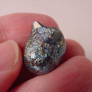 May include: A small, irregularly shaped, iridescent glass bead. The bead has a dark blue base color with silver and gold metallic accents. The surface has a textured, almost crystalline appearance, reflecting light.