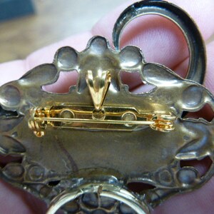 May include: The back of a gold-toned brooch with a detailed design. The brooch has a pin and a loop for hanging.
