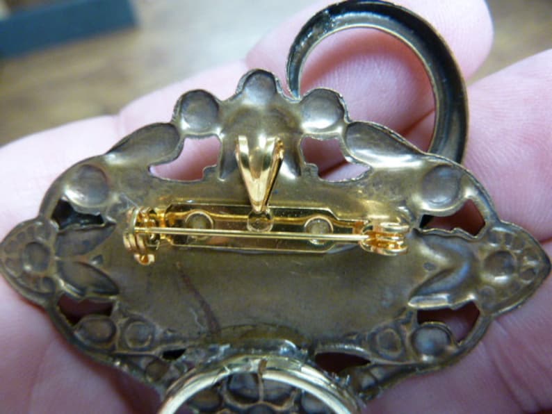 May include: The back of a gold-toned brooch with a detailed design. The brooch has a pin and a loop for hanging.