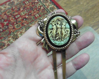May include: A gold hair comb with a decorative oval cameo featuring three women in a green and white design. The cameo is surrounded by a beaded border and a gold bow.