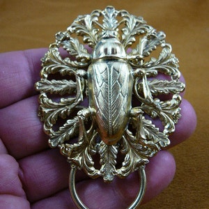 Può includere: Una spilla in metallo dorato con un design di scarabeo dettagliato con intricati accenti floreali. La spilla ha un anello attaccato alla parte inferiore.