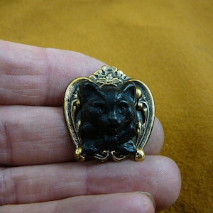 Könnte beinhalten: Eine schwarze Katzenkopf-Brosche mit goldenen Akzenten. Die Katze befindet sich in einem herzförmigen Rahmen mit verzierten Details.